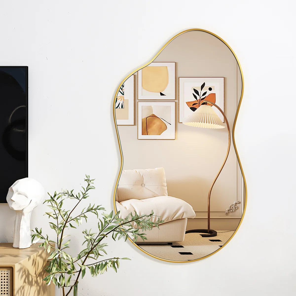 Irregular gold-framed wall mirror with a cloud-like shape reflecting a modern interior featuring a cream tufted armchair, pleated floor lamp, and abstract wall art in warm tones.