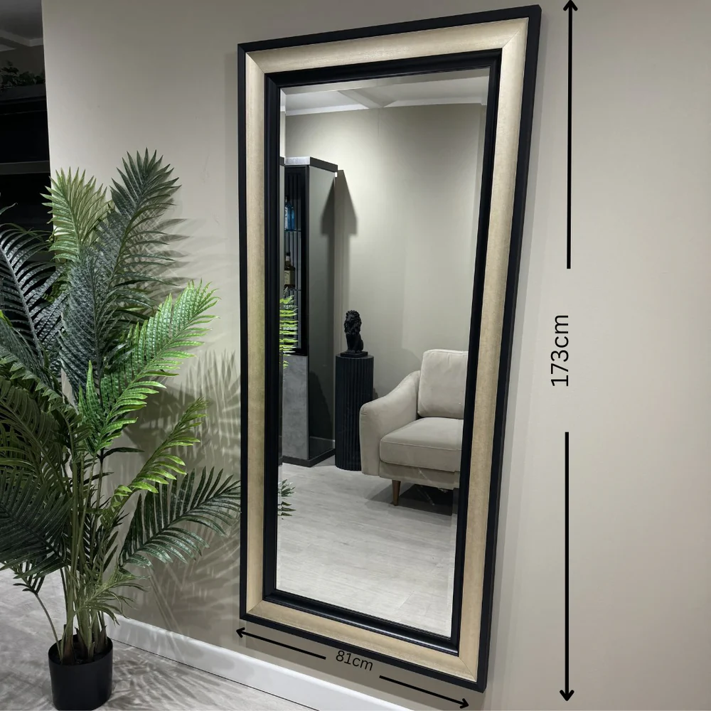 Rectangular wall mirror with a layered black and metallic frame, mounted on a dark blue wall, reflecting a modern interior with light gray walls and wooden flooring.