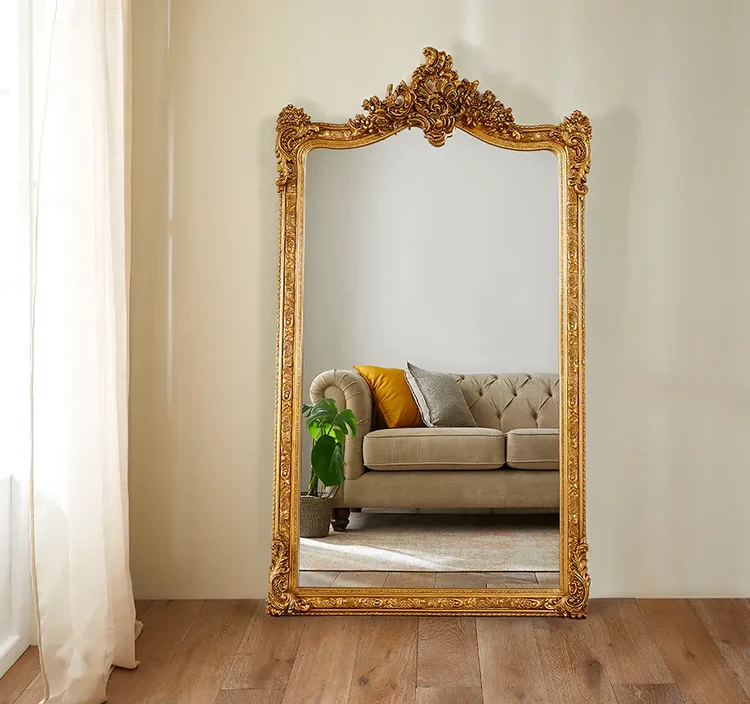 Ornate gold-framed full-length mirror leaning against a wall, reflecting a tufted beige sofa with cushions, a green plant, and a light rug in a cozy living room.