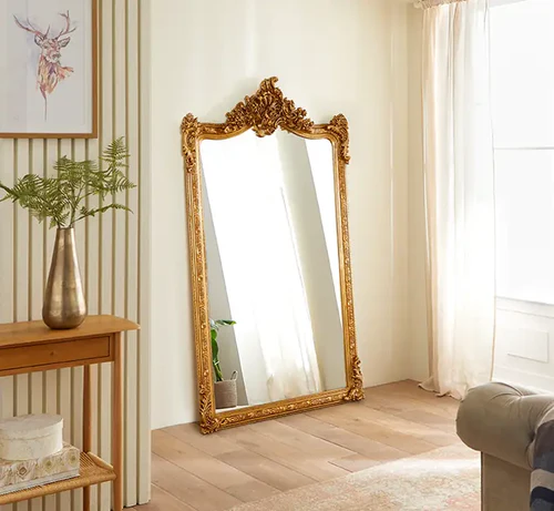 Ornate gold-framed full-length mirror leaning against a wall, reflecting a tufted beige sofa with cushions, a green plant, and a light rug in a cozy living room.