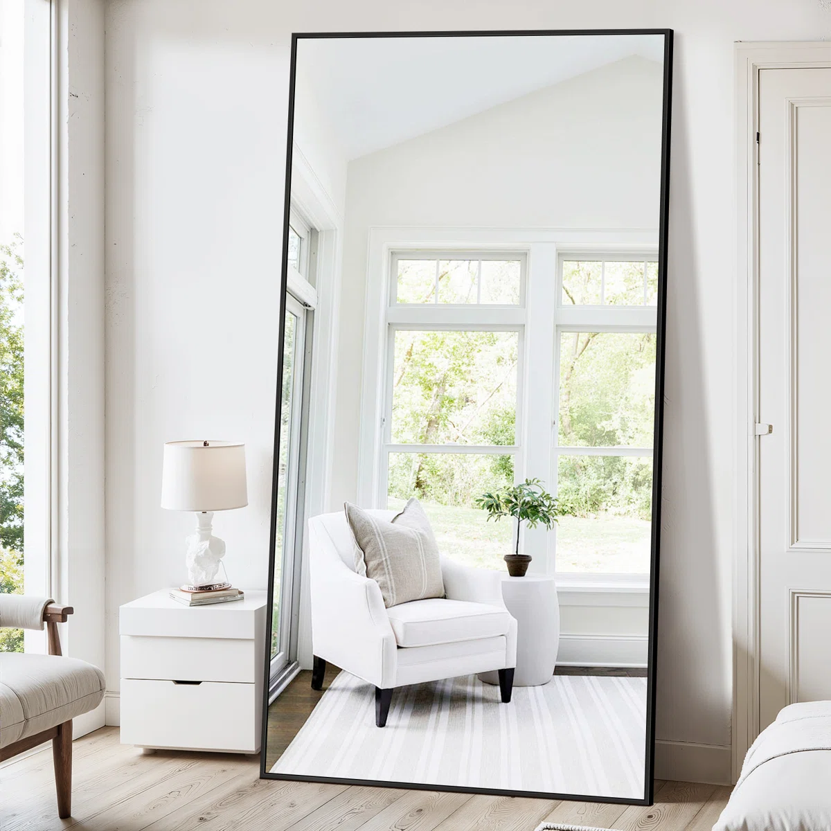 Large mirror reflecting a bright room with white armchair, striped rug, potted plant, and sunlit windows.