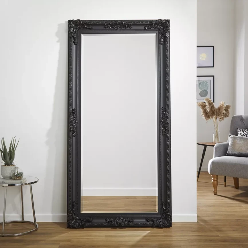 Tall black-framed mirror with ornate carvings leaning against a white wall, reflecting a cozy room with wooden flooring, framed artwork, and a cushioned chair.