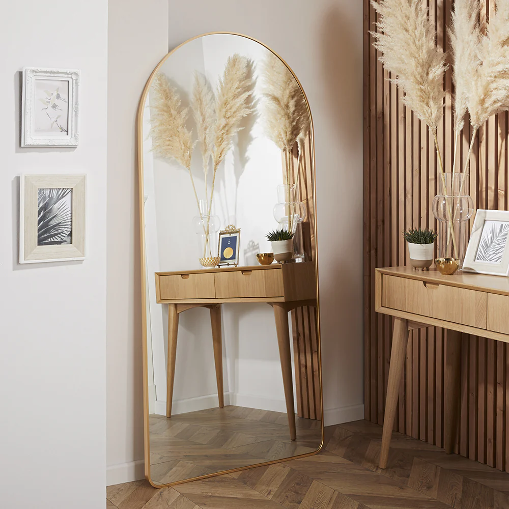 gold-framed mirror reflecting a wooden console table with pampas grass, framed artwork, candles, and a potted plant in a softly lit, modern interior