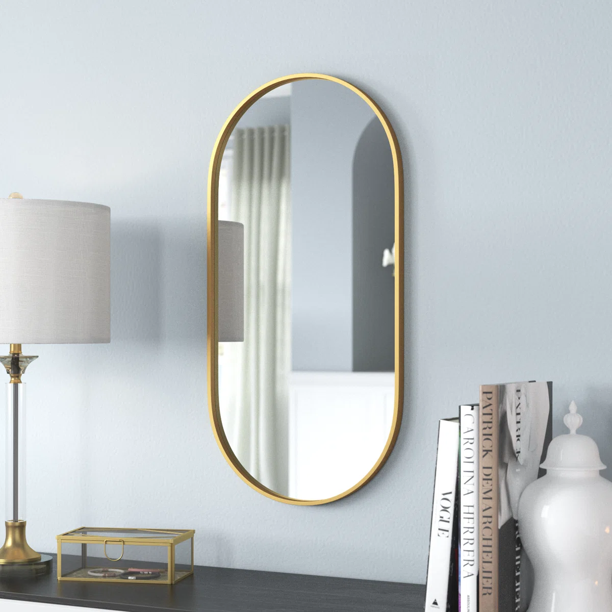 Oval gold-framed wall mirror mounted on a gray wall, reflecting white curtains, arched doorway, and part of a gray lampshade in a modern living space.