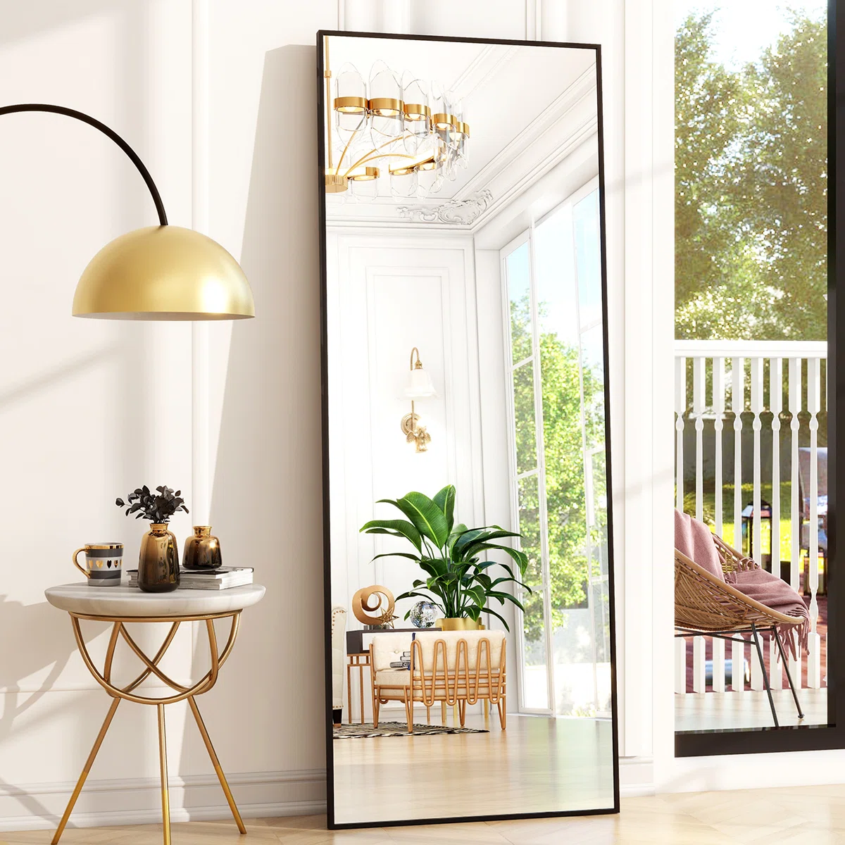 Tall black-framed mirror leaning against a wall, reflecting a bright room with white walls, gold chandelier, green plants, and elegant furniture.