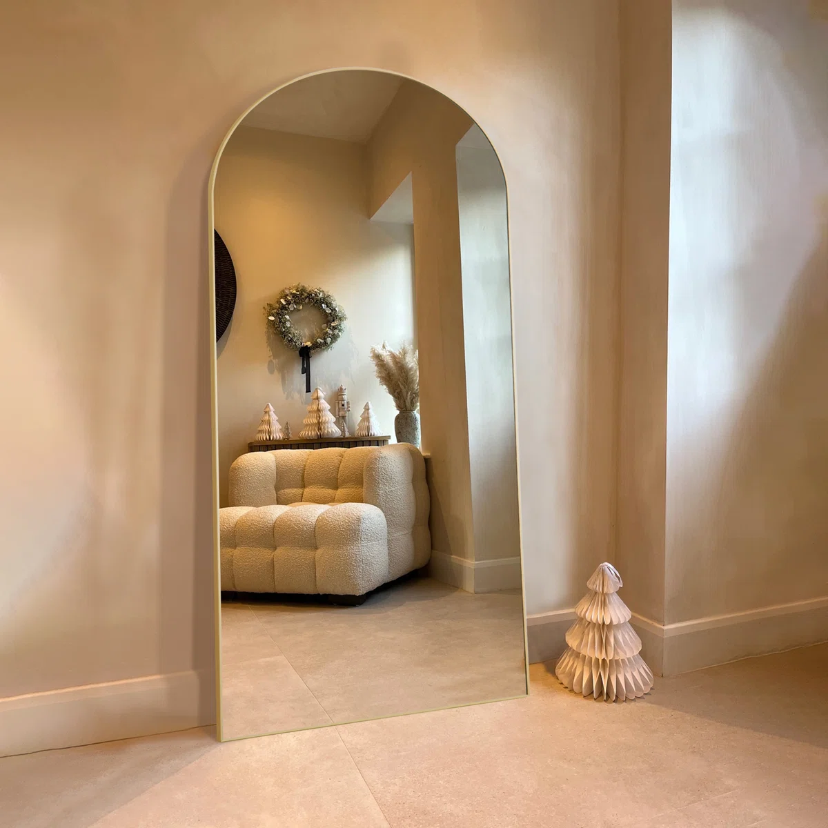 a thin gold frame reflecting a tufted white armchair, pampas grass arrangement, and frosted wreath in a warm, minimalist interior.