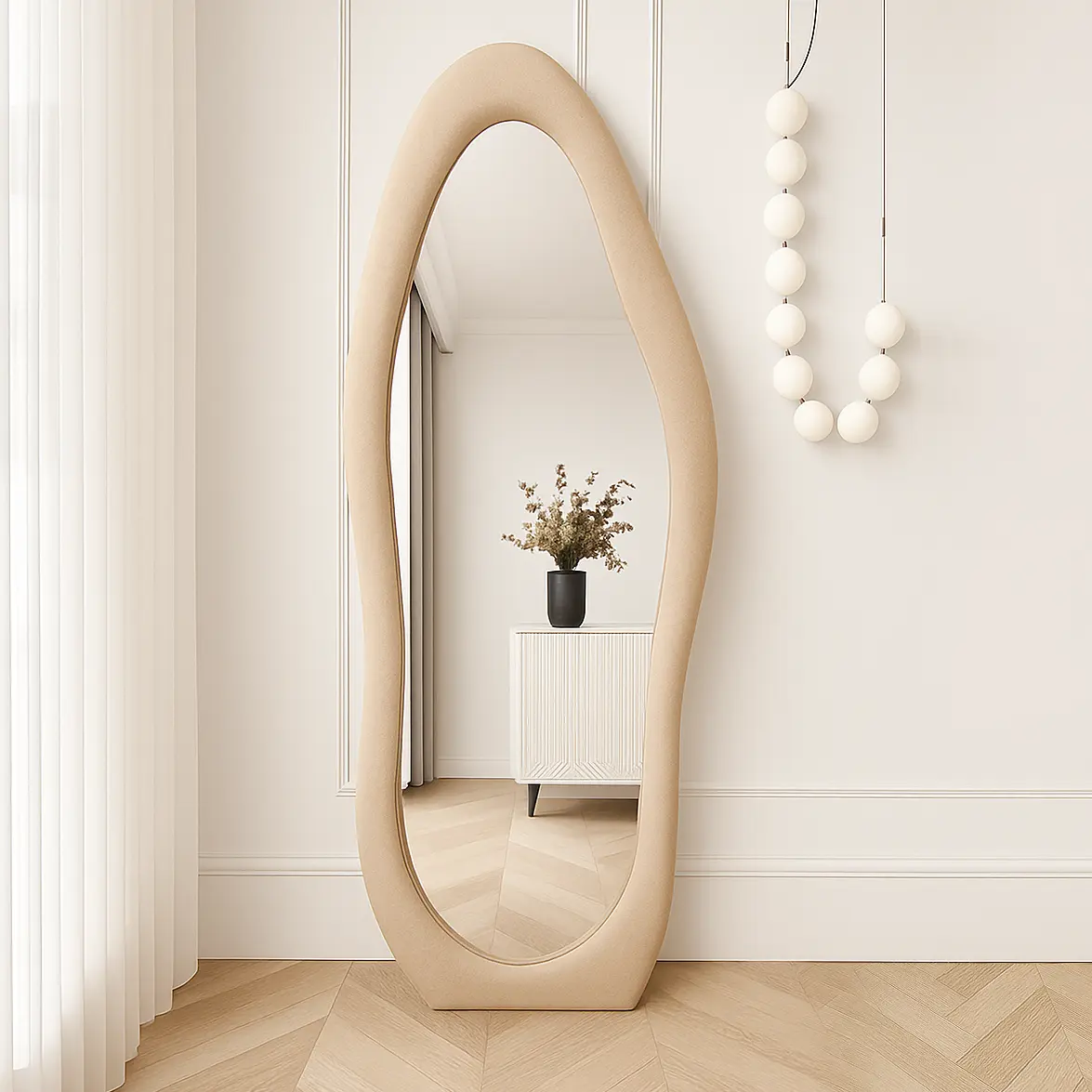Full-length mirror with a beige velvet frame featuring an organic wavy shape, mounted on a white paneled wall beside a decorative hanging and light wood flooring.