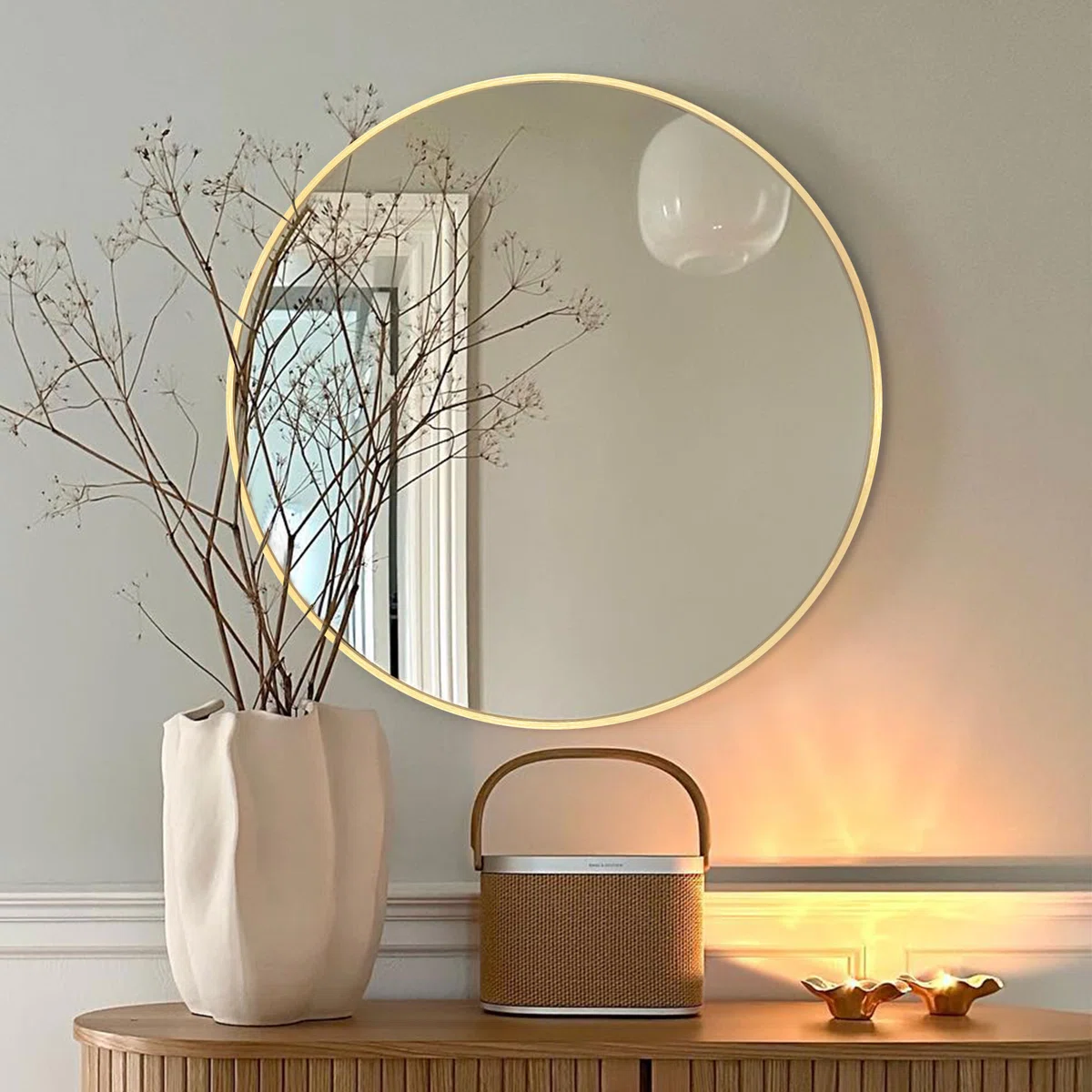 Round wall mirror with thin gold frame reflecting a white door, pendant light, and dried branches against a gray wall.