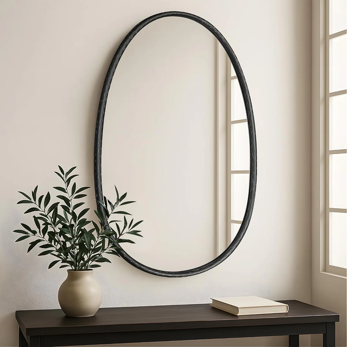 Oval mirror with hammered black metal frame mounted on a light wall, reflecting a window and green plant in a modern interior setting.