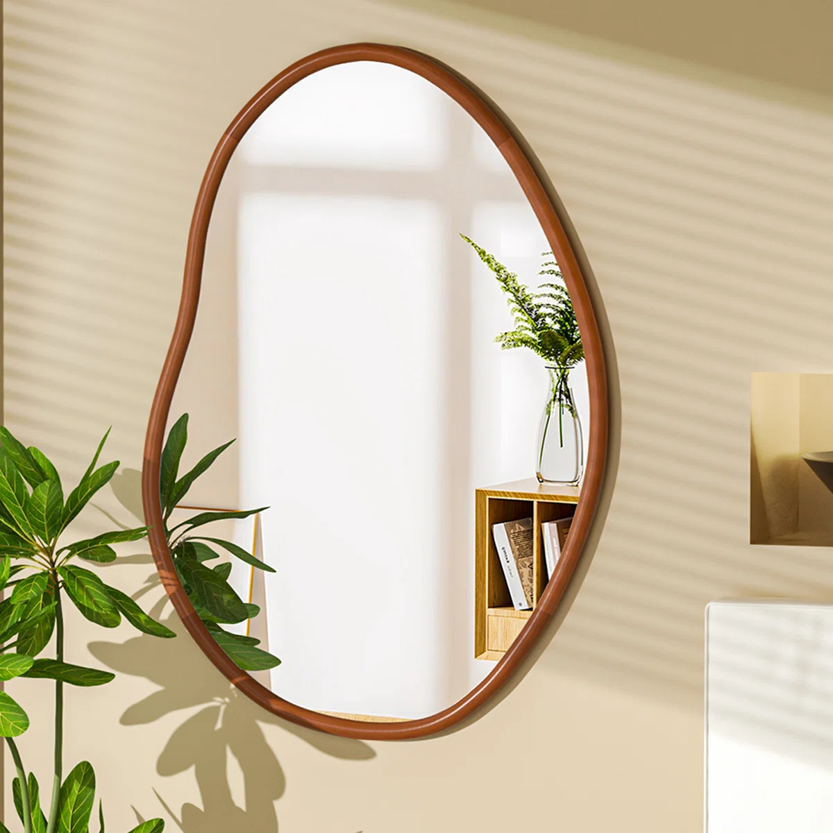 Irregular wall mirror with a beech wood frame mounted on a beige wall, reflecting a sunlit window, a fern in a glass vase, and a wooden shelf with books.