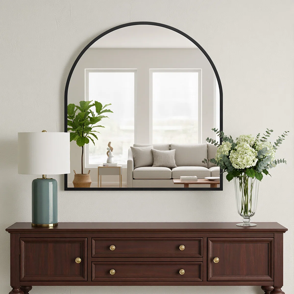 Arched mirror with black frame reflecting a modern living room with gray sofa, wood tables, and sheer curtains.