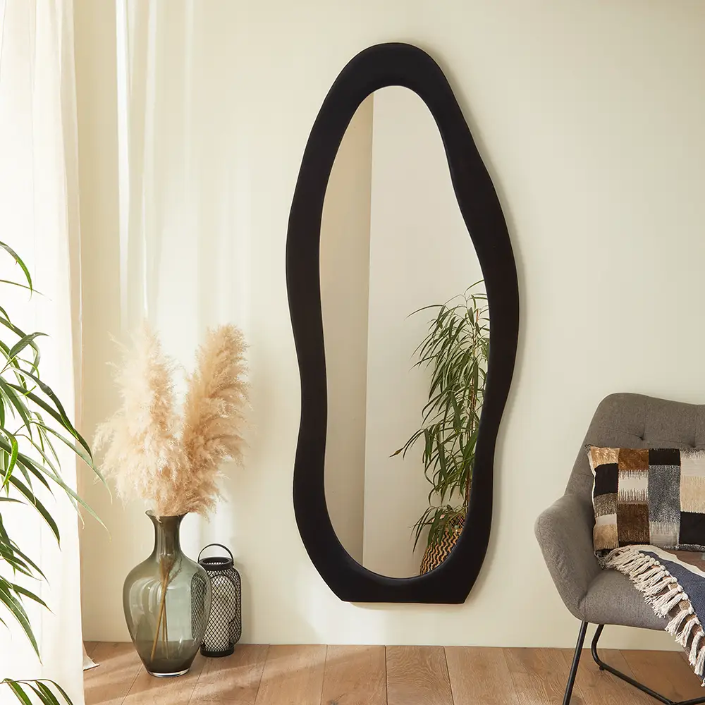 Full-length mirror with a black irregular wavy frame, reflecting a green potted plant, gray chair, and pampas grass in a minimalist interior.