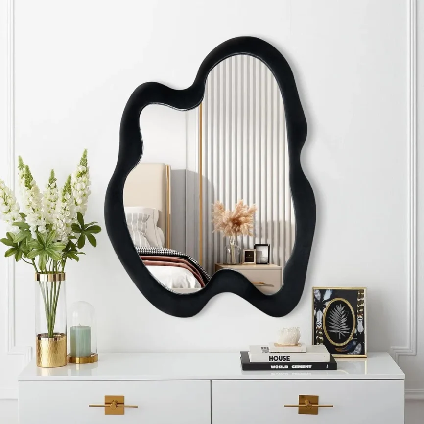 Irregular-shaped wall mirror with black velvet frame mounted on a white wall, reflecting a bedroom with white bedding