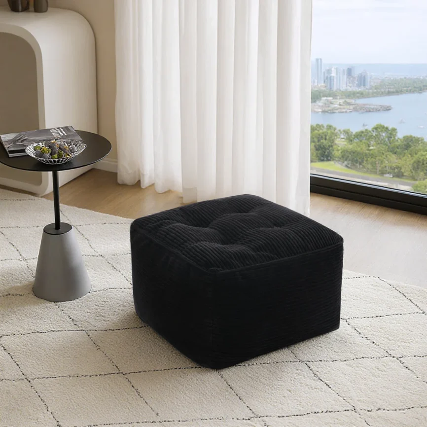 Black square corduroy ottoman with tufted top and stitched panels, placed on a light textured carpet.