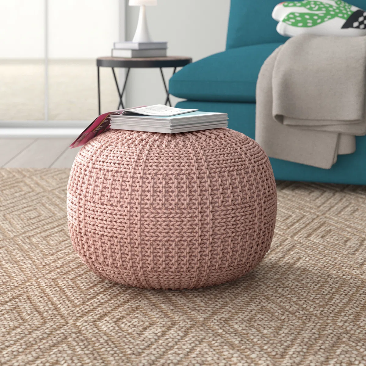 Pink round knitted pouf with square-patterned texture, topped with books and placed on a patterned rug in a cozy living room.