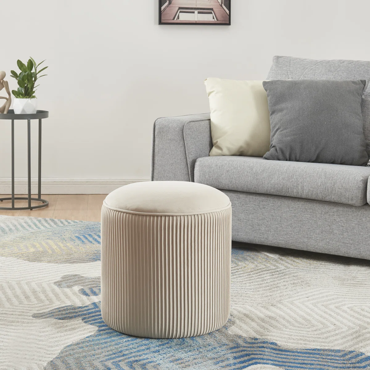 Beige round velvet pouf with ribbed sides and cushioned top, placed beside a gray sofa on a patterned rug.