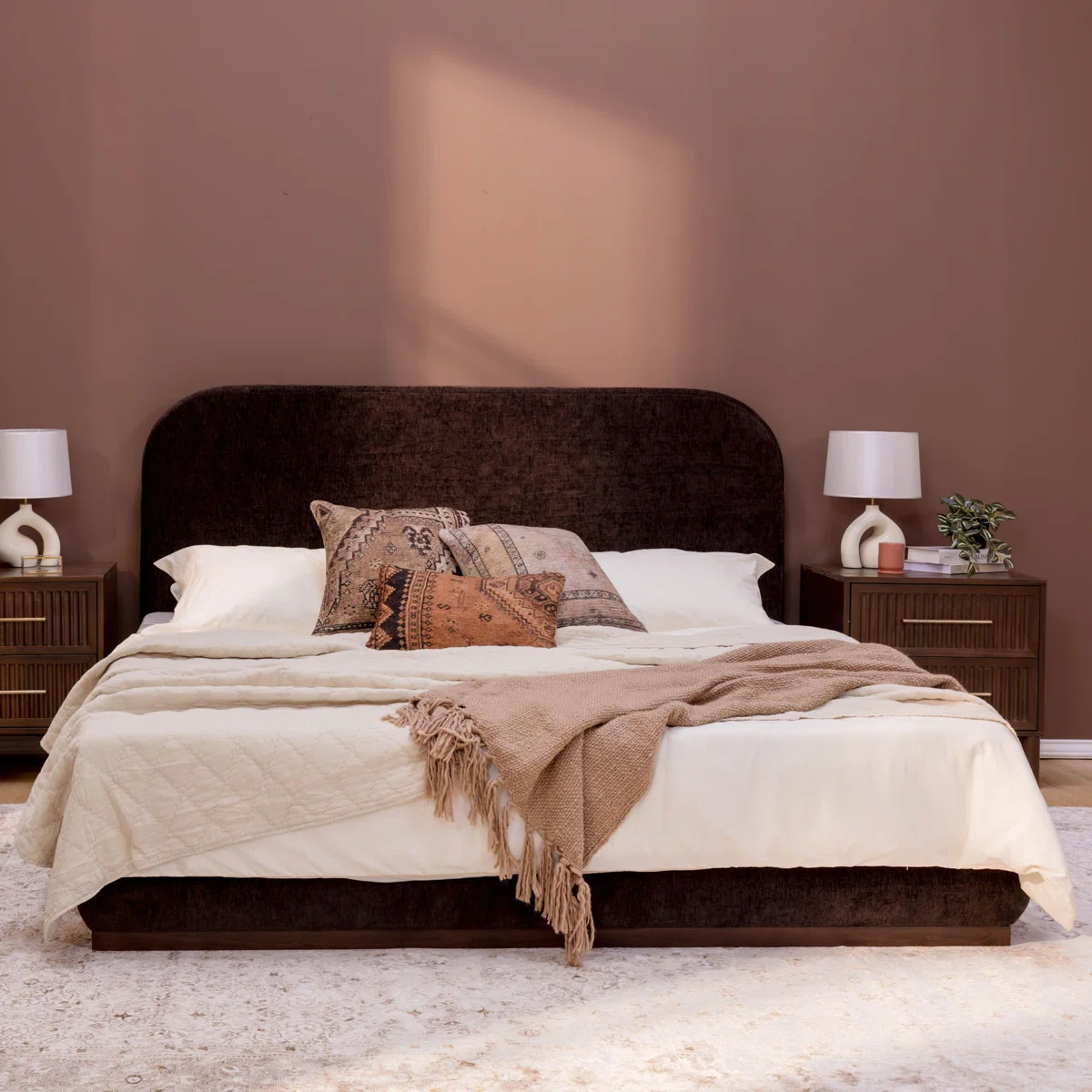 A king-size bed with a dark upholstered headboard, styled in crisp white bedding and layered pillows in neutral tones.
